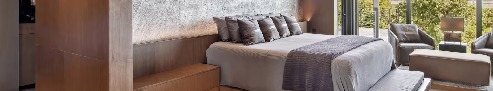 Modern bedroom with a large gray-upholstered bed, wooden accents, and glass doors opening to a green outdoor view.